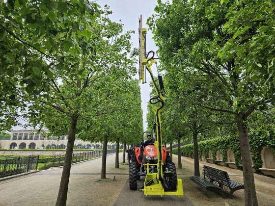 A red tractor with a tall yellow shearing device that is being used to trim trees. 
